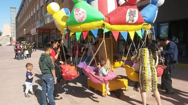 新疆，強烈反差的一幕：孩子們在玩旋轉木馬，在他們的身後不遠處，和田市場「後方護衛隊」正在演練。