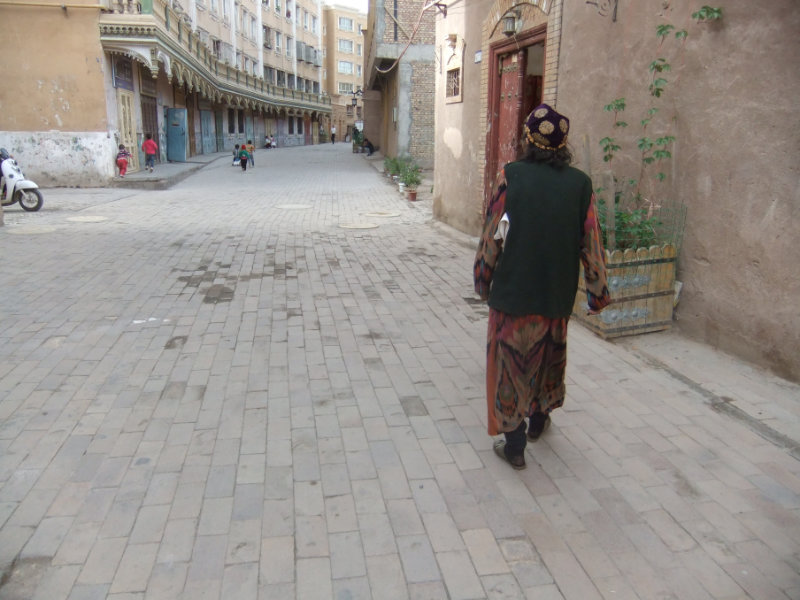 一名老年維吾爾族婦女正走在重建、拓寬後的大路上。現在諷刺的是她不得不戴維吾爾族花帽，而不是她的頭巾。這種帽子一般是年輕女孩或婦女的打扮，而頭巾當下則被視為忠實於「極端」伊斯蘭教的象徵。喀什　2018年